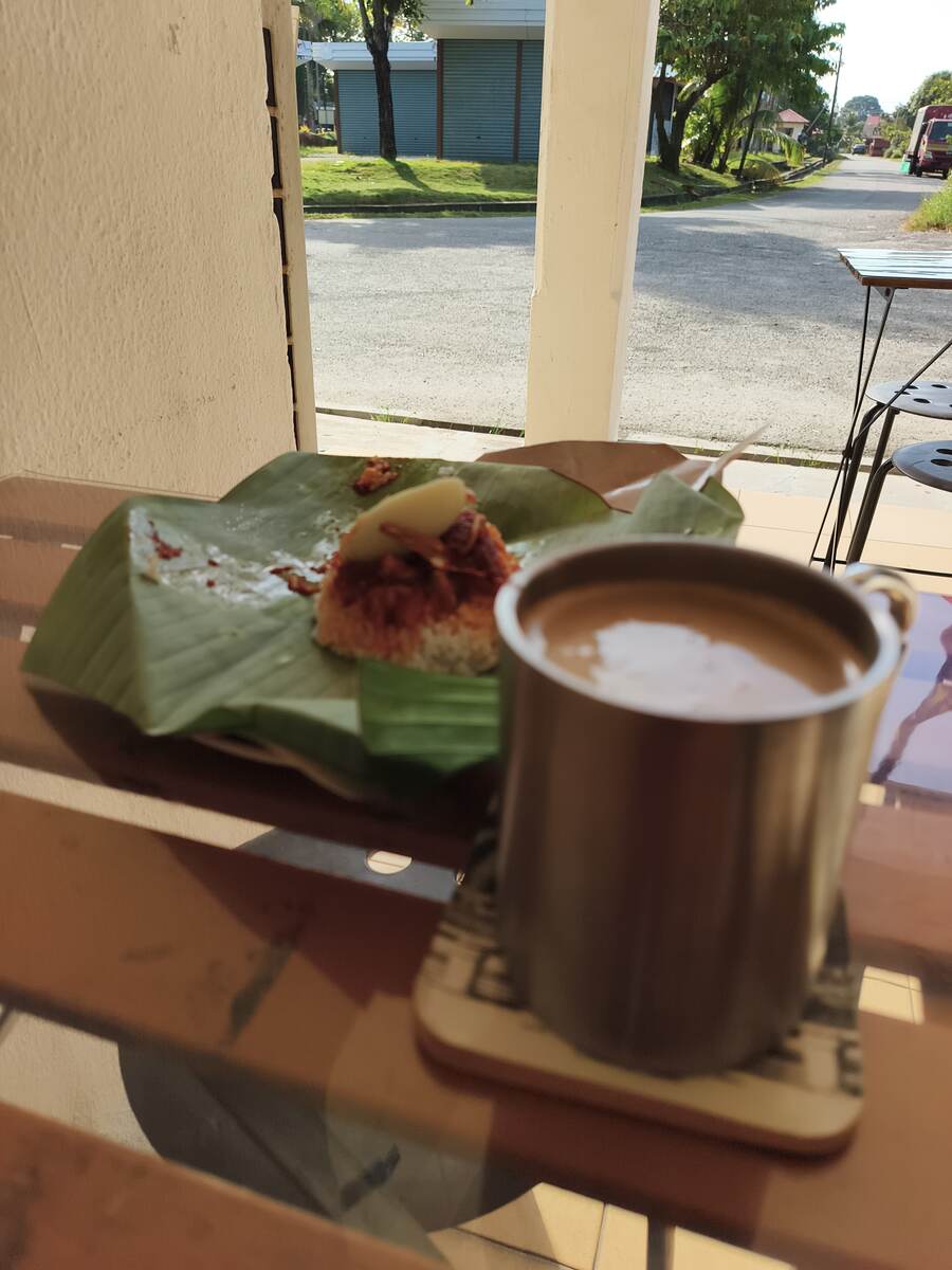 Nasi lemak on banana leaf beside hot kopi in a stainless camping mug at the cafe's roadside table